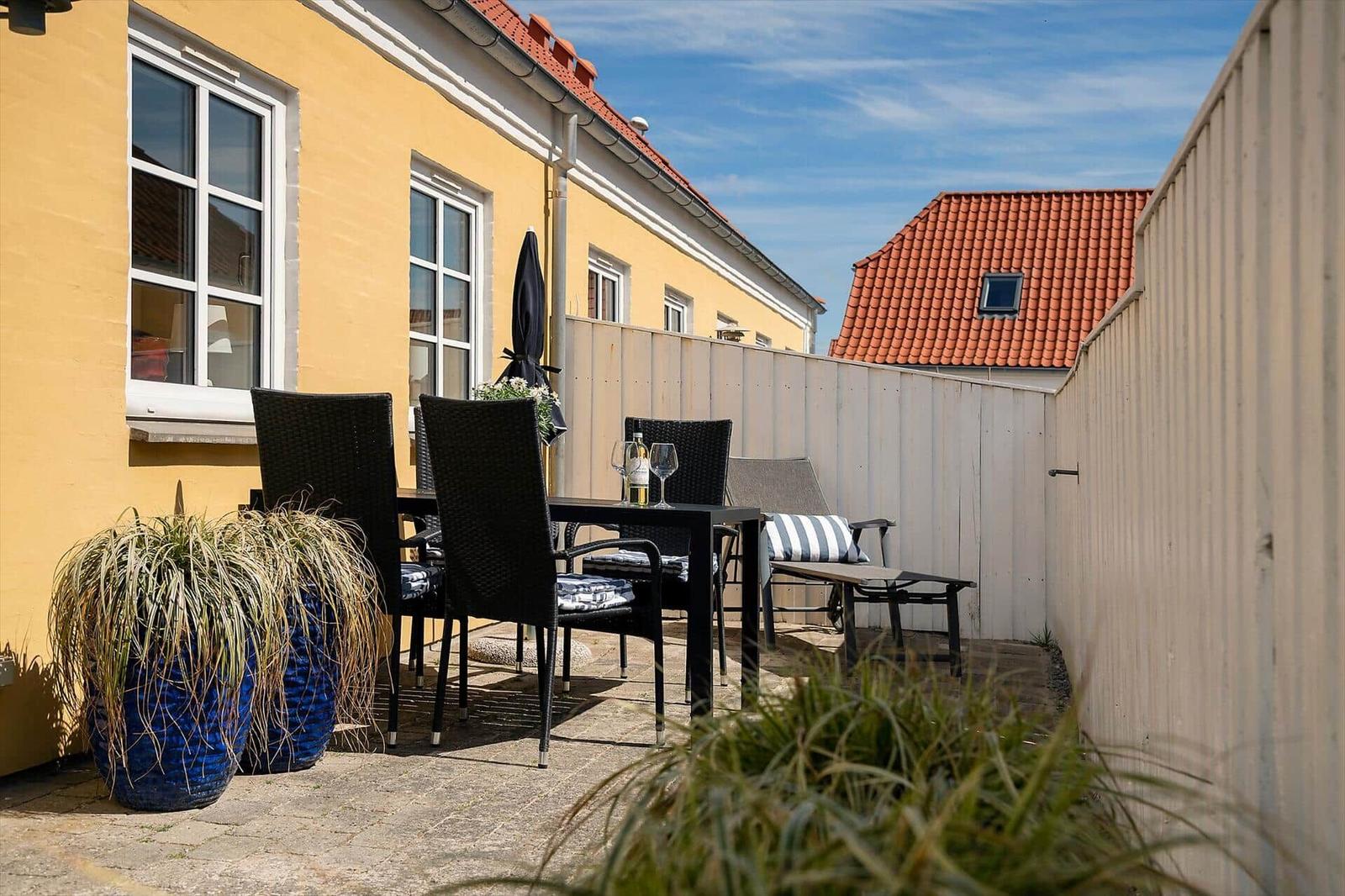 Terrasse mit Tisch, Stühlen und Pflanzen an gelbem Haus mit rotem Dach.