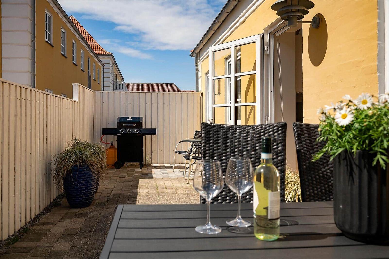 Terrasse mit Tisch, Stühlen, Grill und Blumen. Hinterhof mit gelbem Haus.