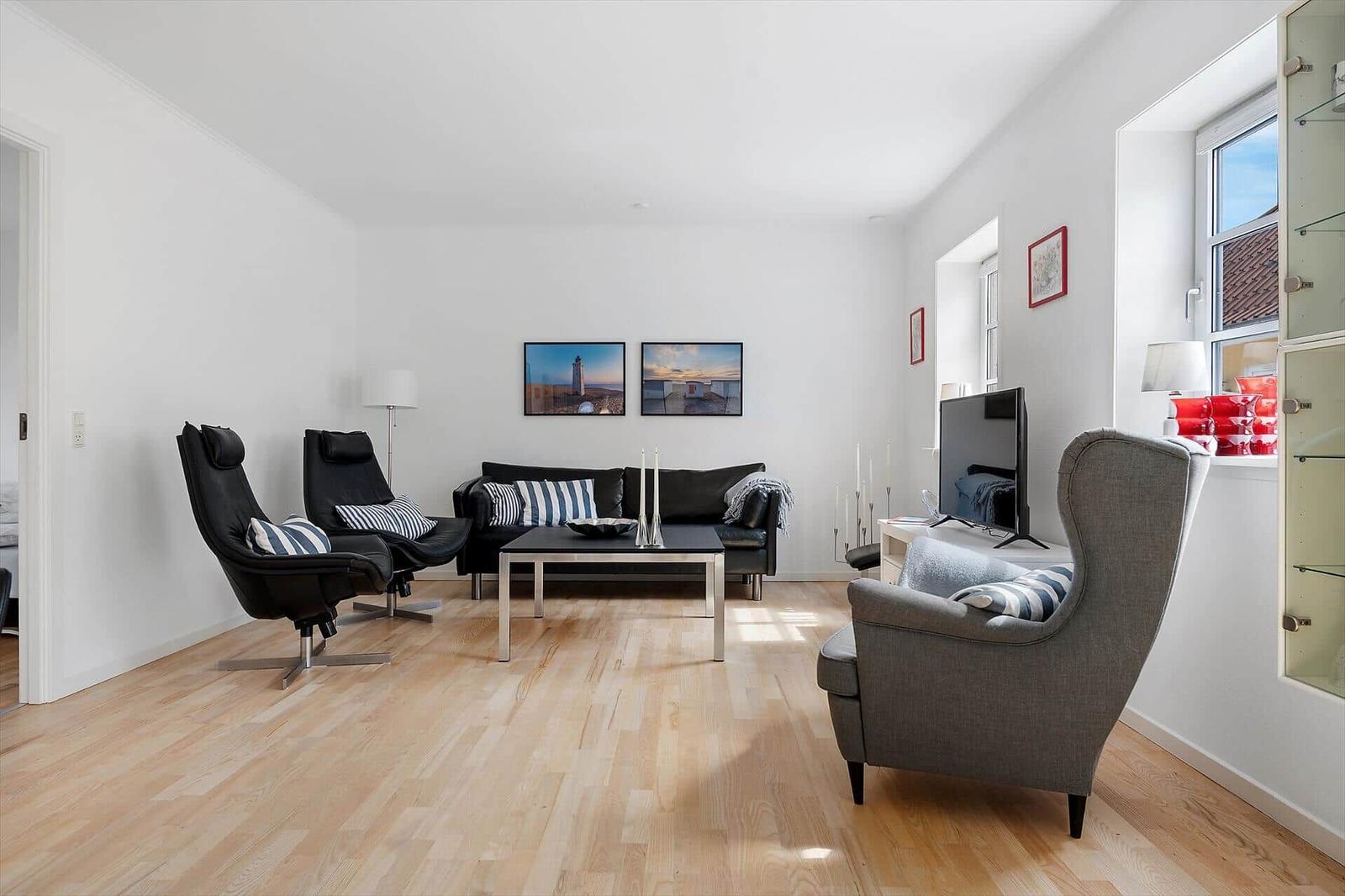 Wohnzimmer mit schwarzer Couch, zwei Sesseln und Holzboden. An der Wand hängen zwei Bilder.