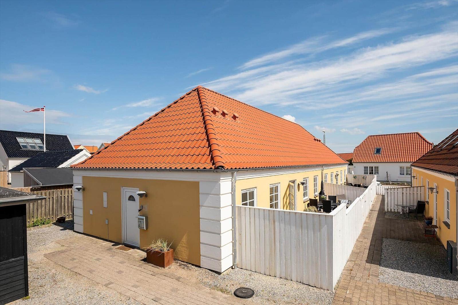 Haus mit rotem Dach und gelber Fassade, umgeben von weißem Zaun und gepflastertem Hof.
