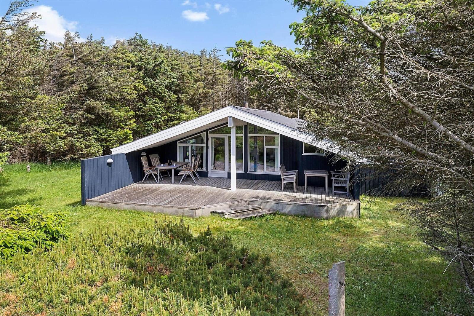 Ein Haus mit Holzterrasse und Garten. Umgeben von Bäumen unter blauem Himmel.