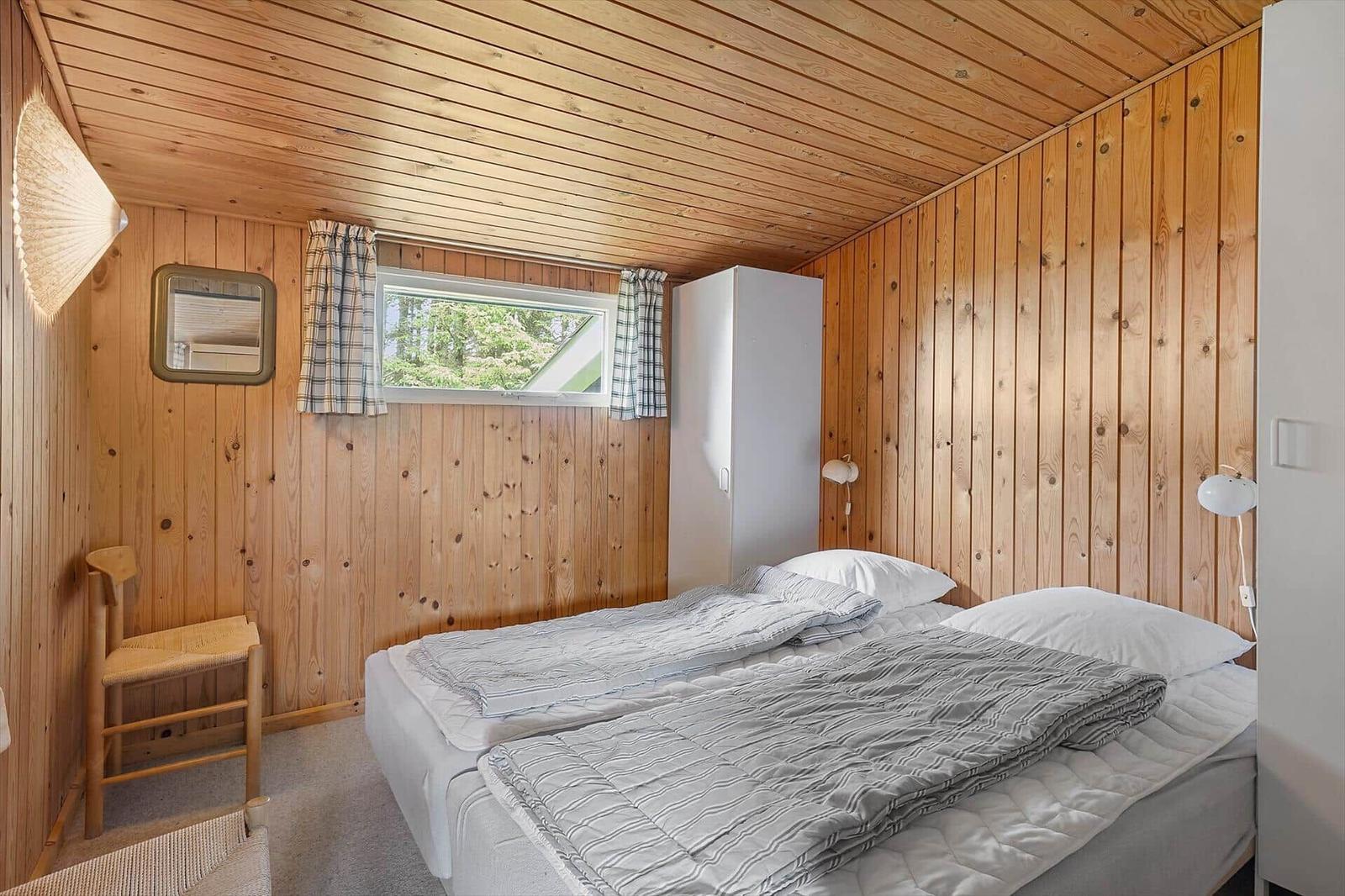 Schlafzimmer mit Holzwand, Bett, Kleiderschrank und Fenster mit Blick auf Wald.