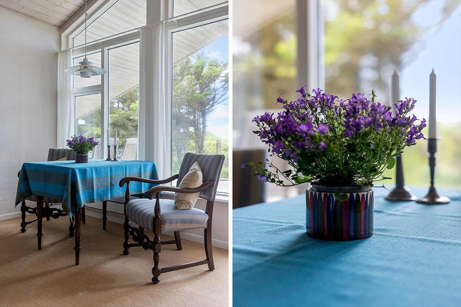 Esstisch mit blauer Tischdecke und Stuhl vor großem Fenster. Vase mit lila Blumen und Kerzenhaltern.