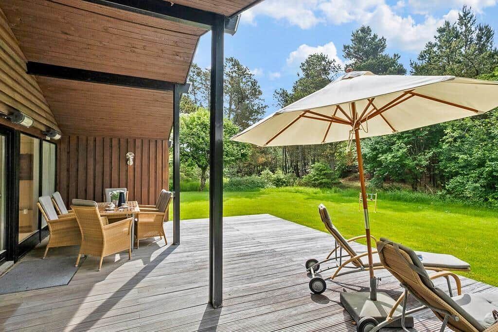 Deck with table, chairs, umbrella, and loungers before green lawn and forest.