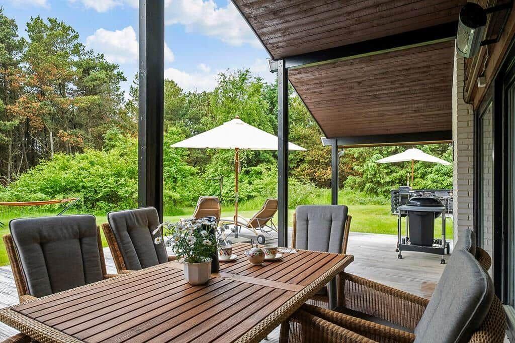 Terrace with dining table, chairs, umbrellas, and grill. View of green landscape.