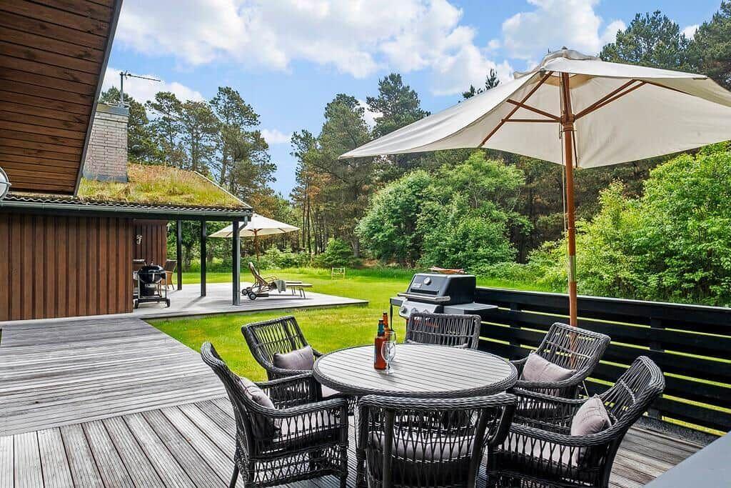 Deck with table, chairs, and umbrella. Grill and forest in background.