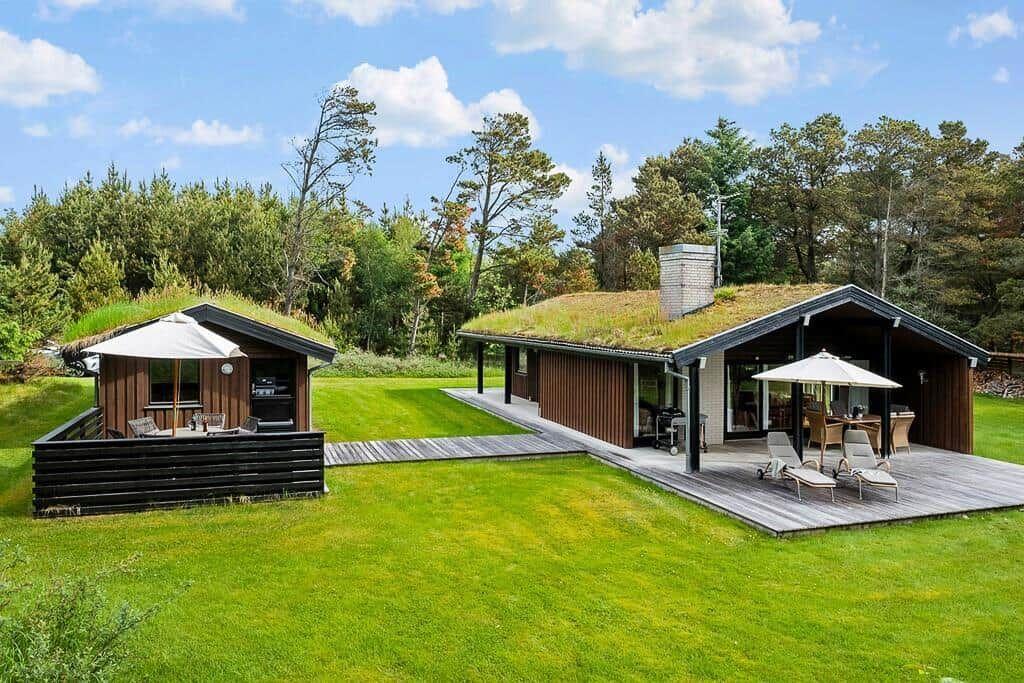 Two houses with grass roofs stand on green lawn. Terraces with umbrellas and lounge chairs.