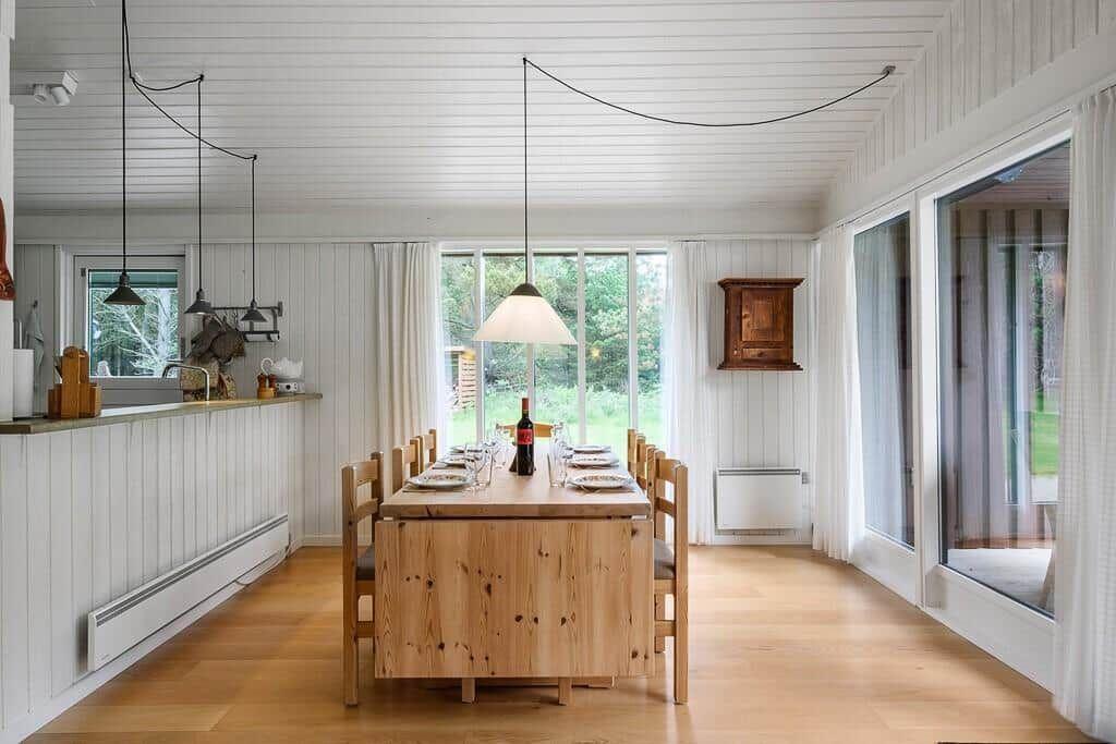 Dining room with wooden table, chairs, and windows to the green outdoor area.