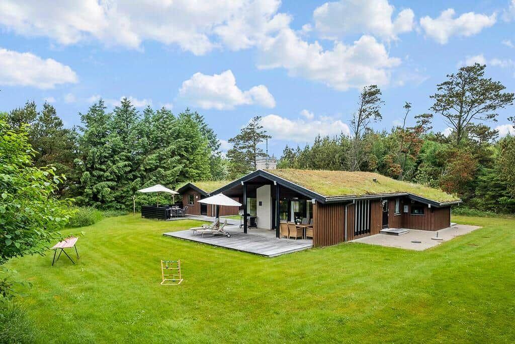 Modern wooden cabin with grass roof, terrace, and garden. Surrounded by trees under blue sky.