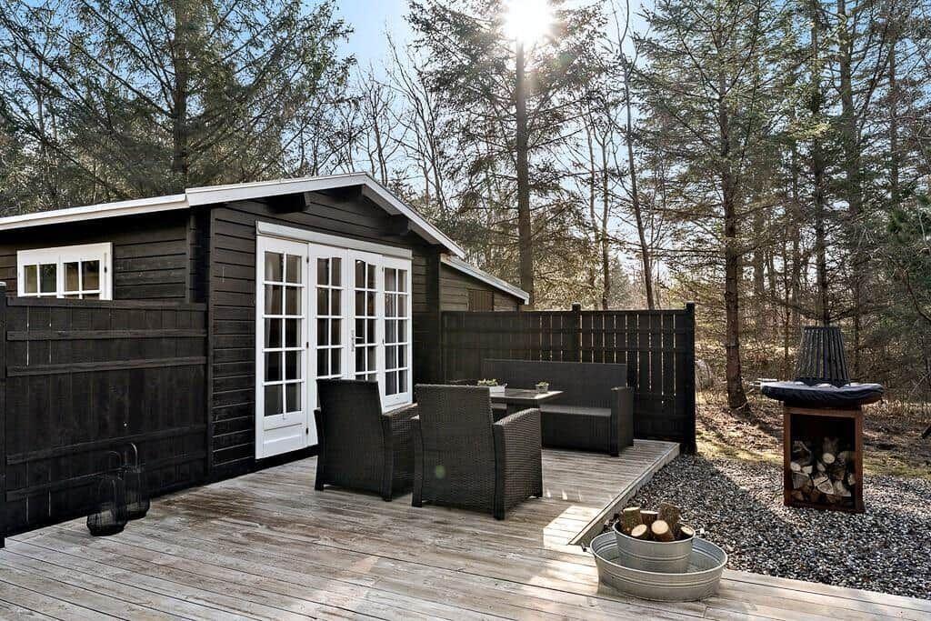 Schwarze Holzhütte mit Terrasse, Sitzgruppe und Feuerkorb im Wald.