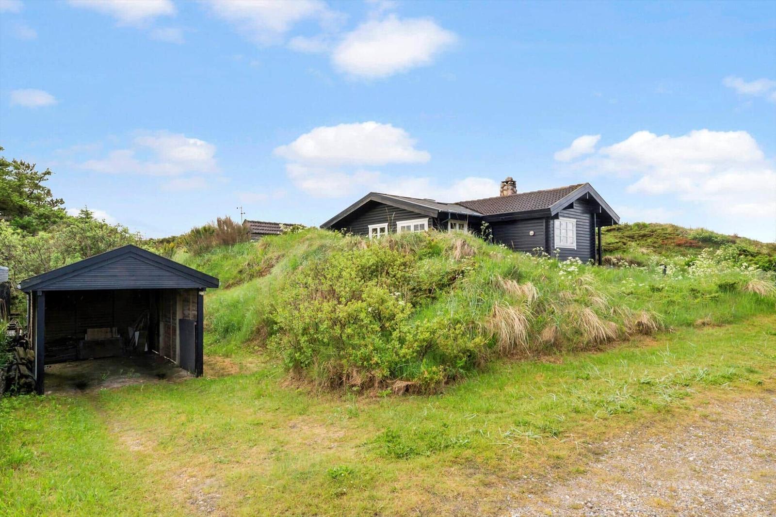 Ein dunkles Holzhaus mit Dach und Schornstein liegt auf einer grünen Anhöhe. Links steht eine Garage.
