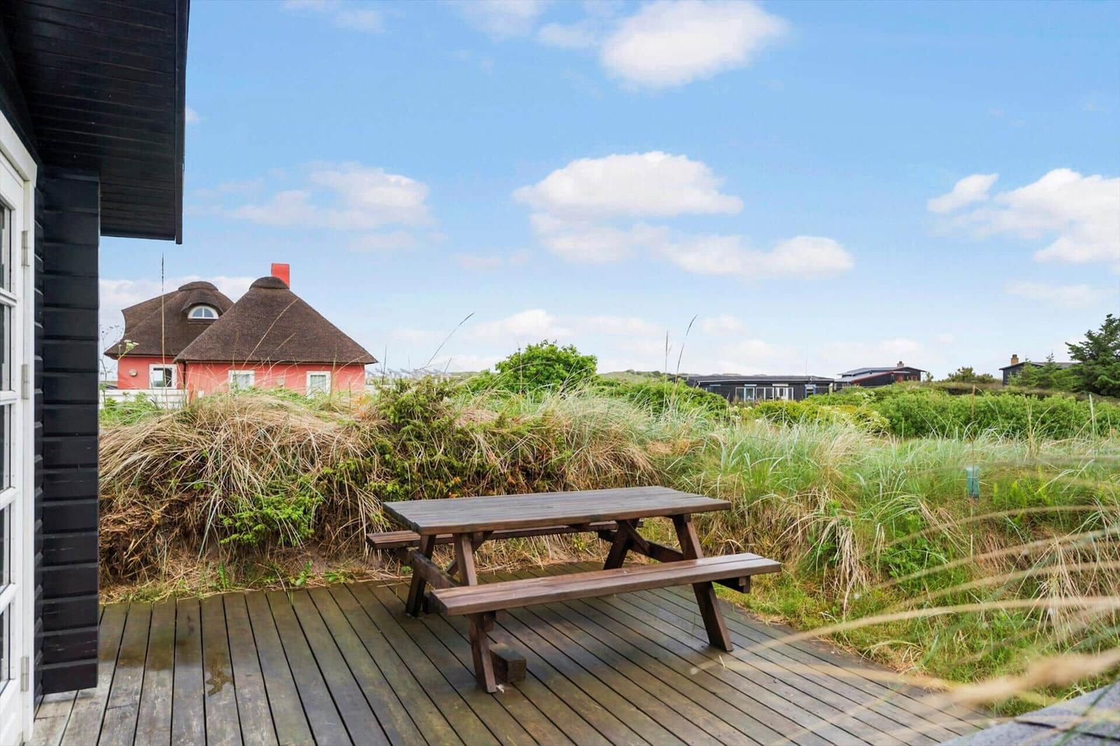 Holzterrasse mit Picknicktisch. Blick auf Grasland und ein rotes Haus mit Strohdach.