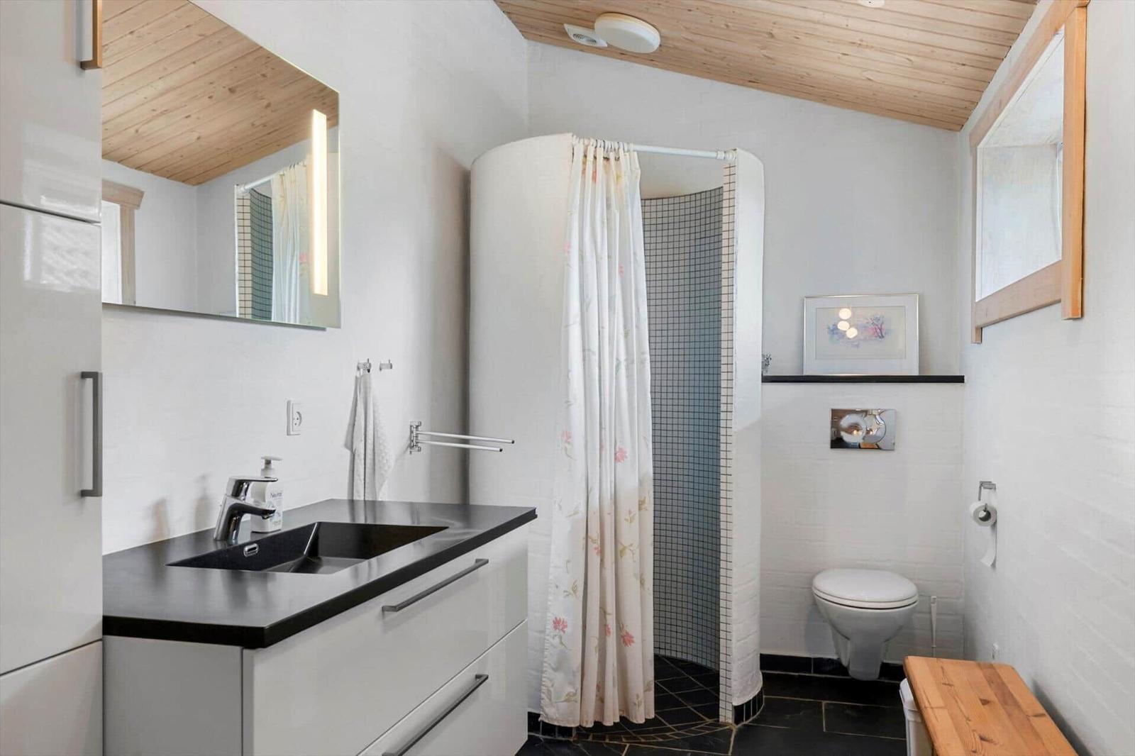 Badezimmer mit Dusche, Waschbecken und Toilette. Holzdecke und schwarzer Boden.