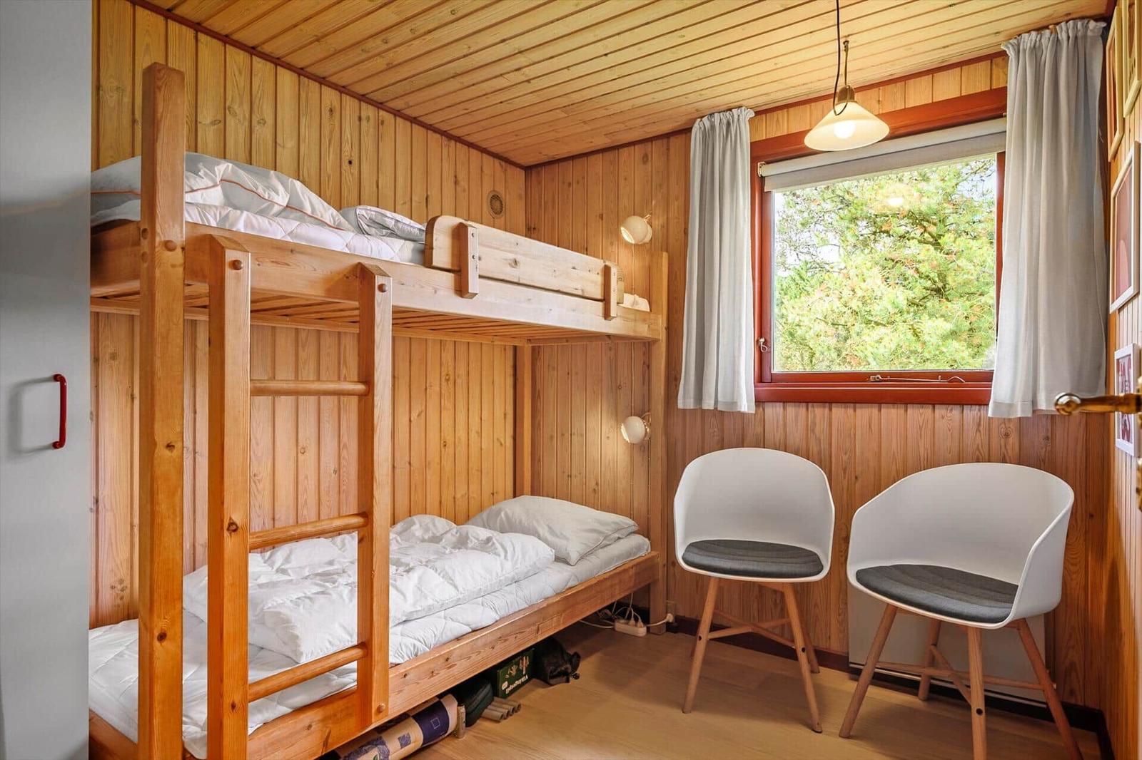Zimmer mit Holzwand, Doppelstockbett und zwei weißen Stühlen neben Fenster.