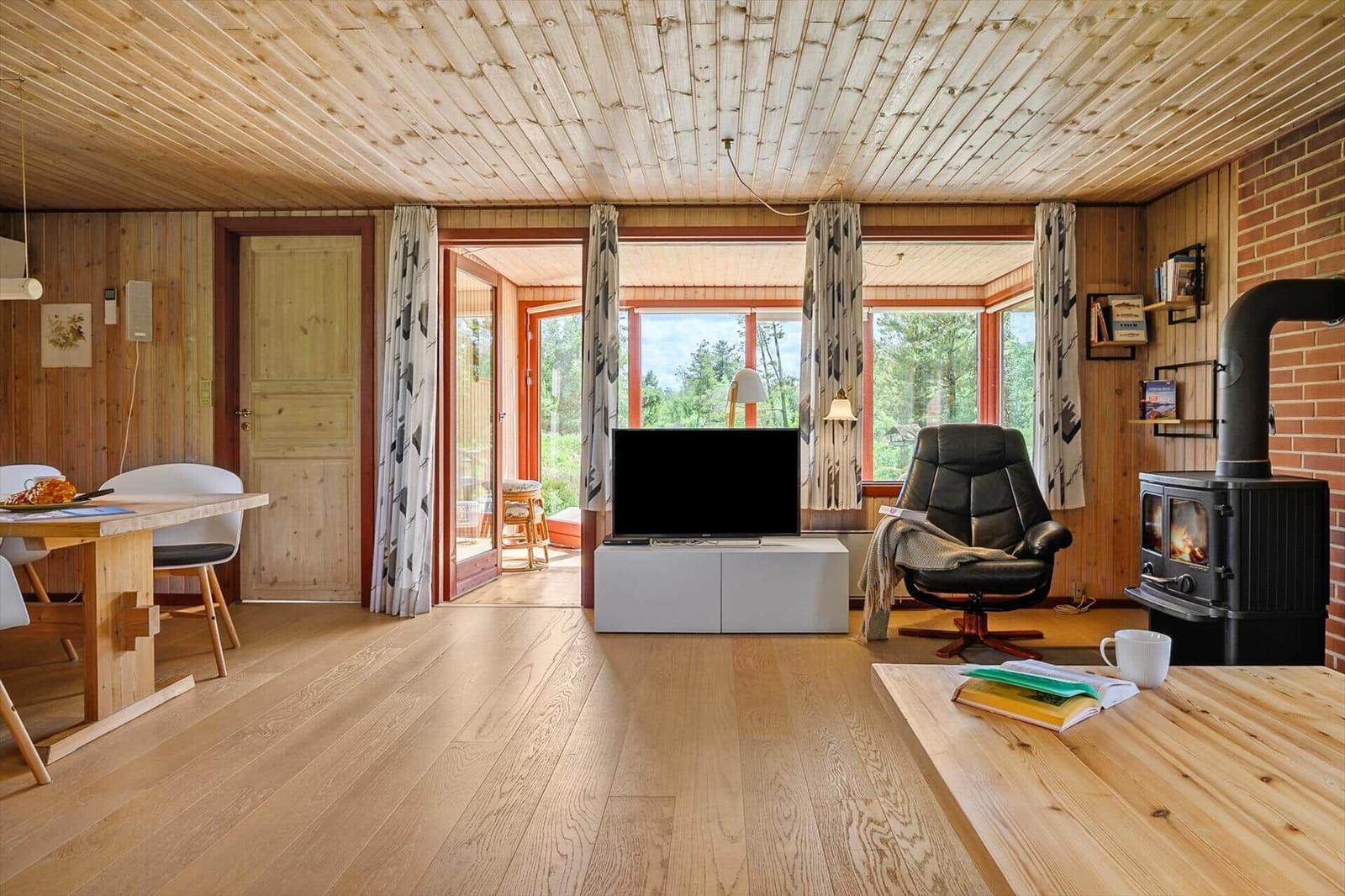 Wohnzimmer mit Holzdecke, TV, Sessel und Kaminofen. Durchgang zur Terrasse mit Blick ins Grüne.