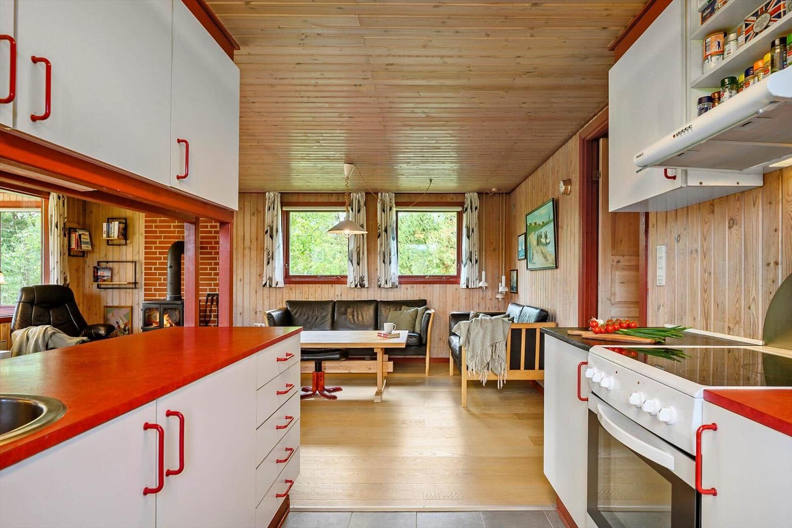 Küche mit rotem Arbeitsplatz und weißer Einbauküche. Wohnbereich mit Sofa und Fenstern.