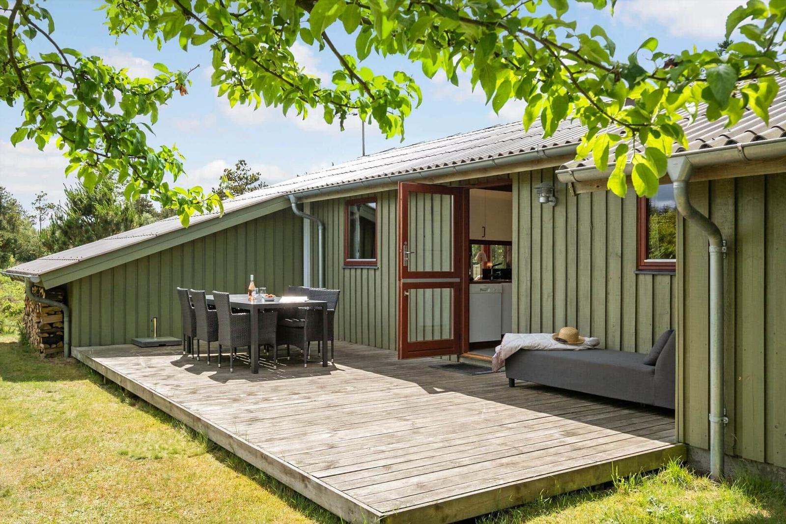 Holzterrasse mit Tisch, Stühlen und Liege vor einem Haus mit offener Tür.