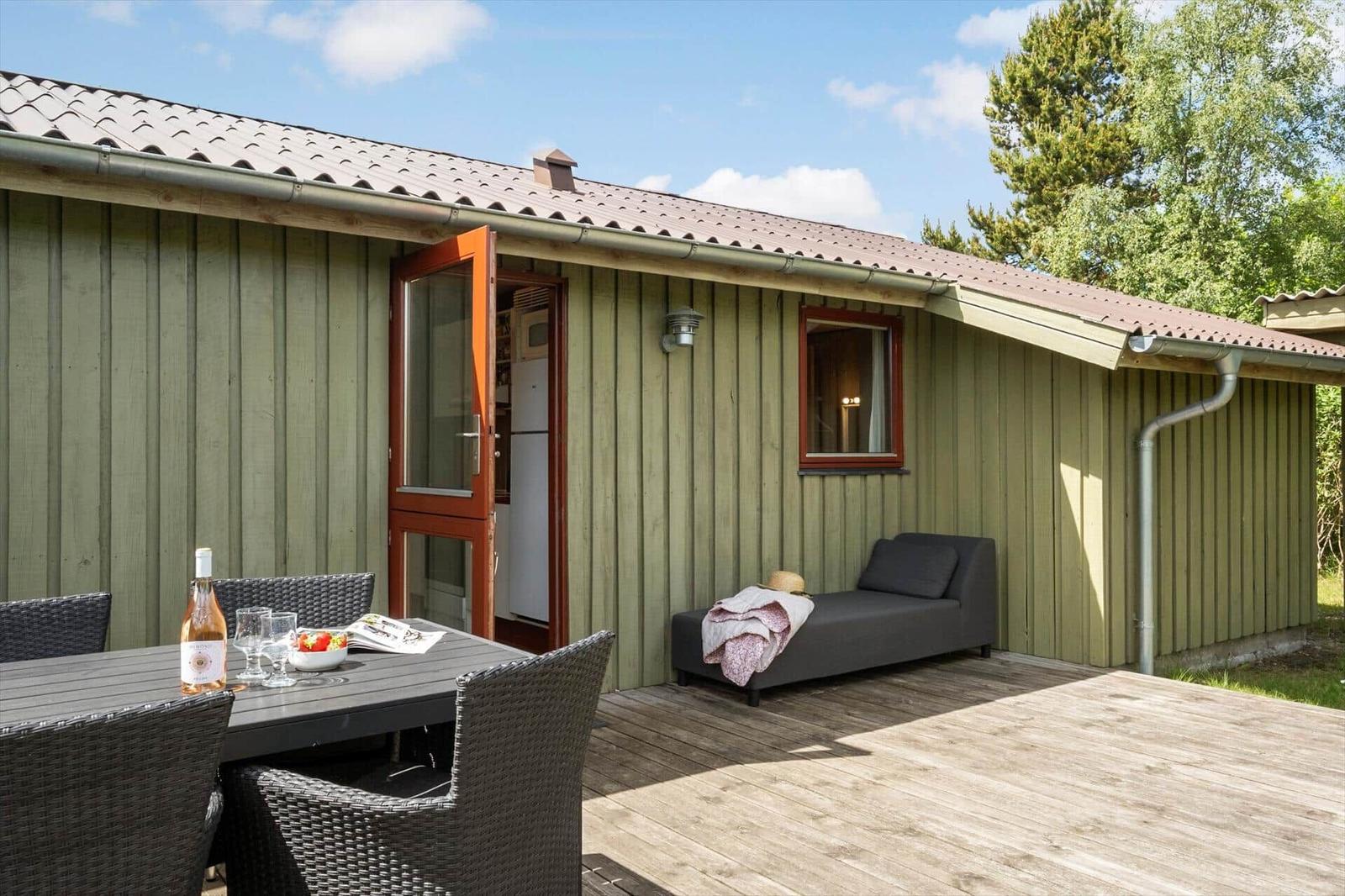 Terrasse mit Tisch, Stühlen und Liege. Hintergrund: Holzhaus mit offener Tür.
