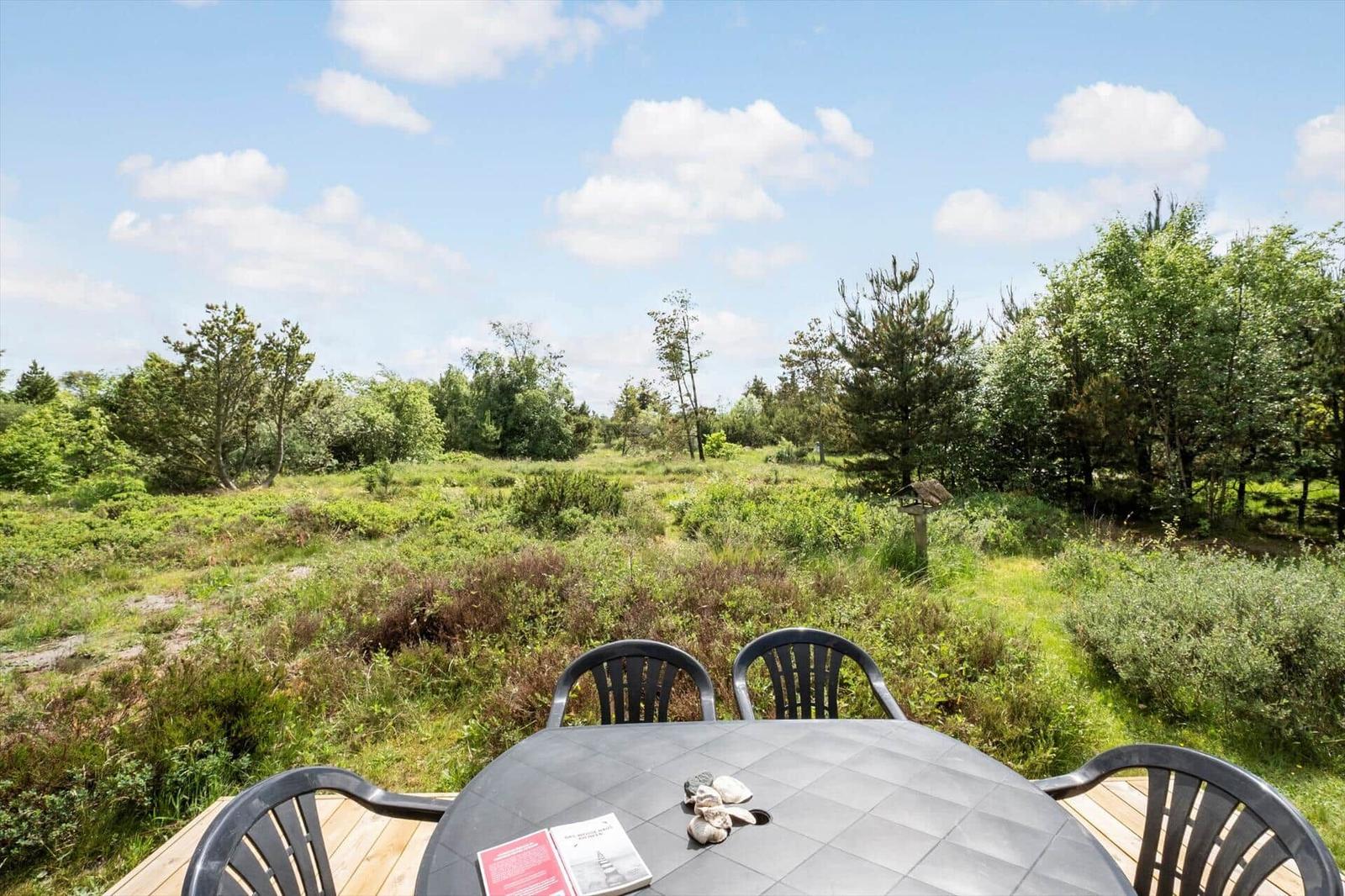 Rundtisch mit Stühlen auf Terrasse, Blick auf grüne Wiese und Bäume unter blauem Himmel.