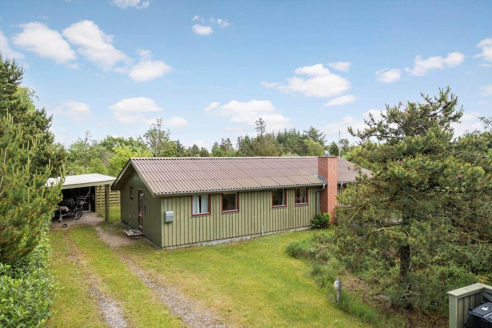 Ein grünes Haus mit braunem Dach steht auf einem Grasgrundstück. Umgeben von Bäumen und einem kleinen Carport.