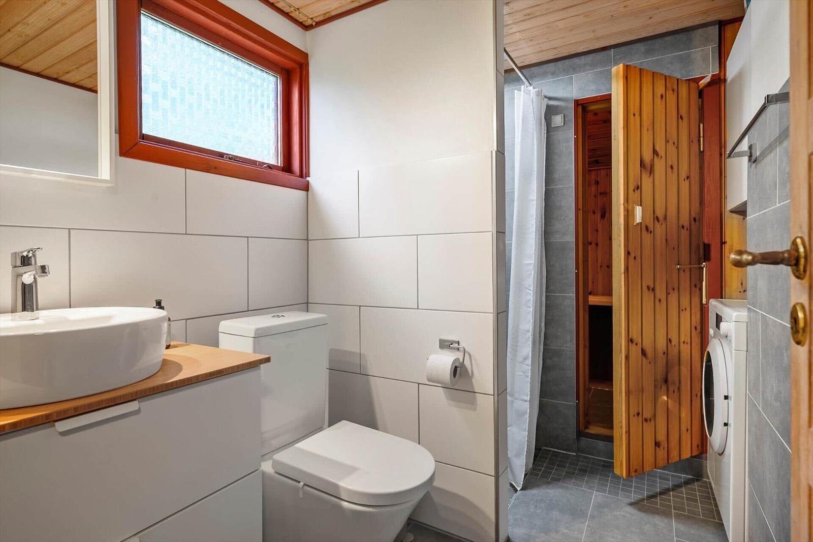 Badezimmer mit Waschbecken, Toilette und Dusche. Holzfenster und Sauna sind sichtbar.