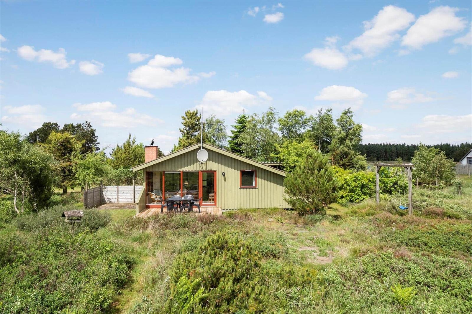 Ein kleines Haus mit Terrasse, umgeben von Grün und Bäumen unter blauem Himmel.