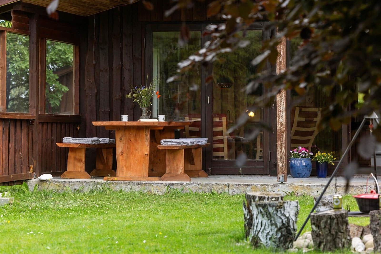 Holztisch mit Bänken auf Terrasse vor Holzhaus mit Blumen und Pflanzen.