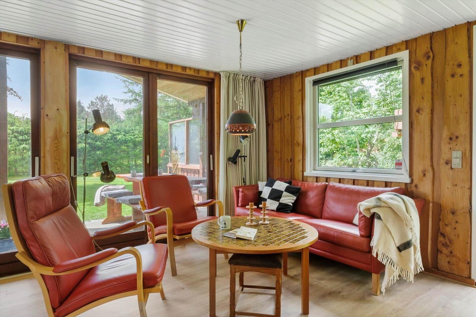 Ein Wohnzimmer mit roten Ledermöbeln, Holzwänden und großen Fenstern zum Garten.