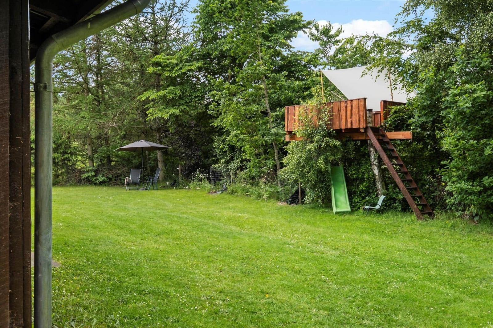 Grüner Garten mit Baumhaus, Rutsche und Sonnenschirm.