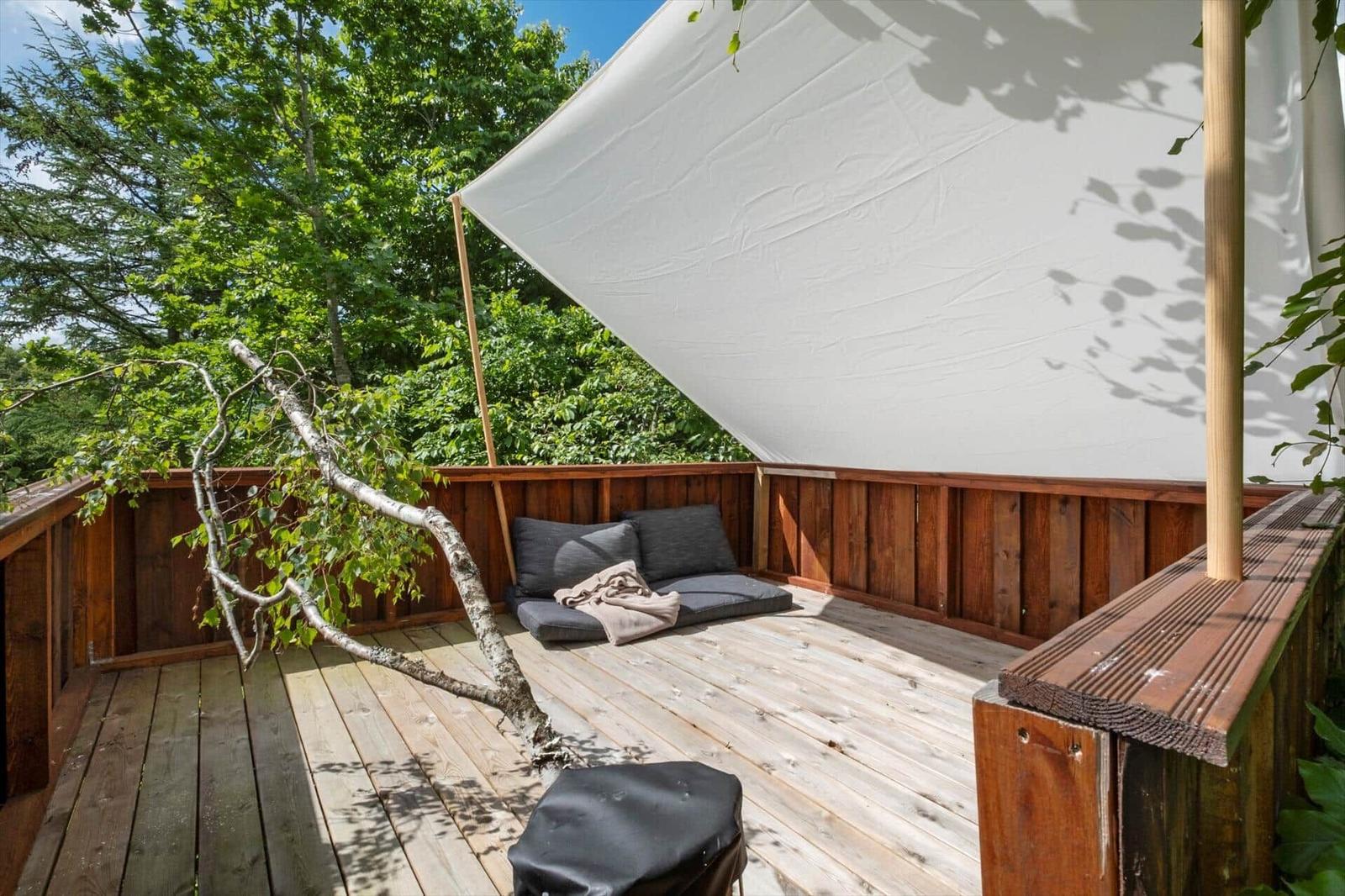 Holzterrasse mit Sitzbank, Sonnensegel und Baumast. Umgeben von Bäumen.