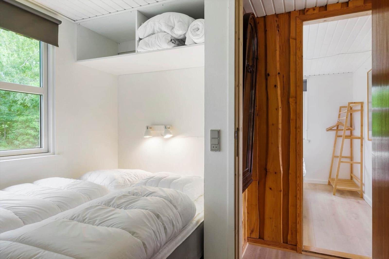 Schlafzimmer mit Bett, Fenster und Aufbewahrung. Holztür führt in einen Flur mit Kleiderständer.