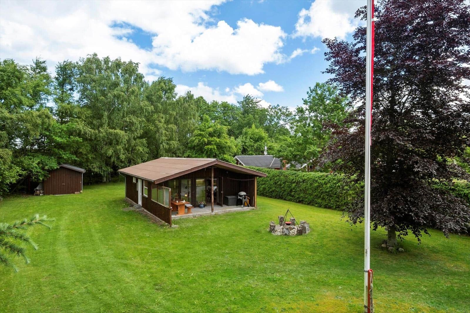 Holzhaus mit Terrasse, Grill und Feuerstelle auf grünem Grundstück umgeben von Bäumen.