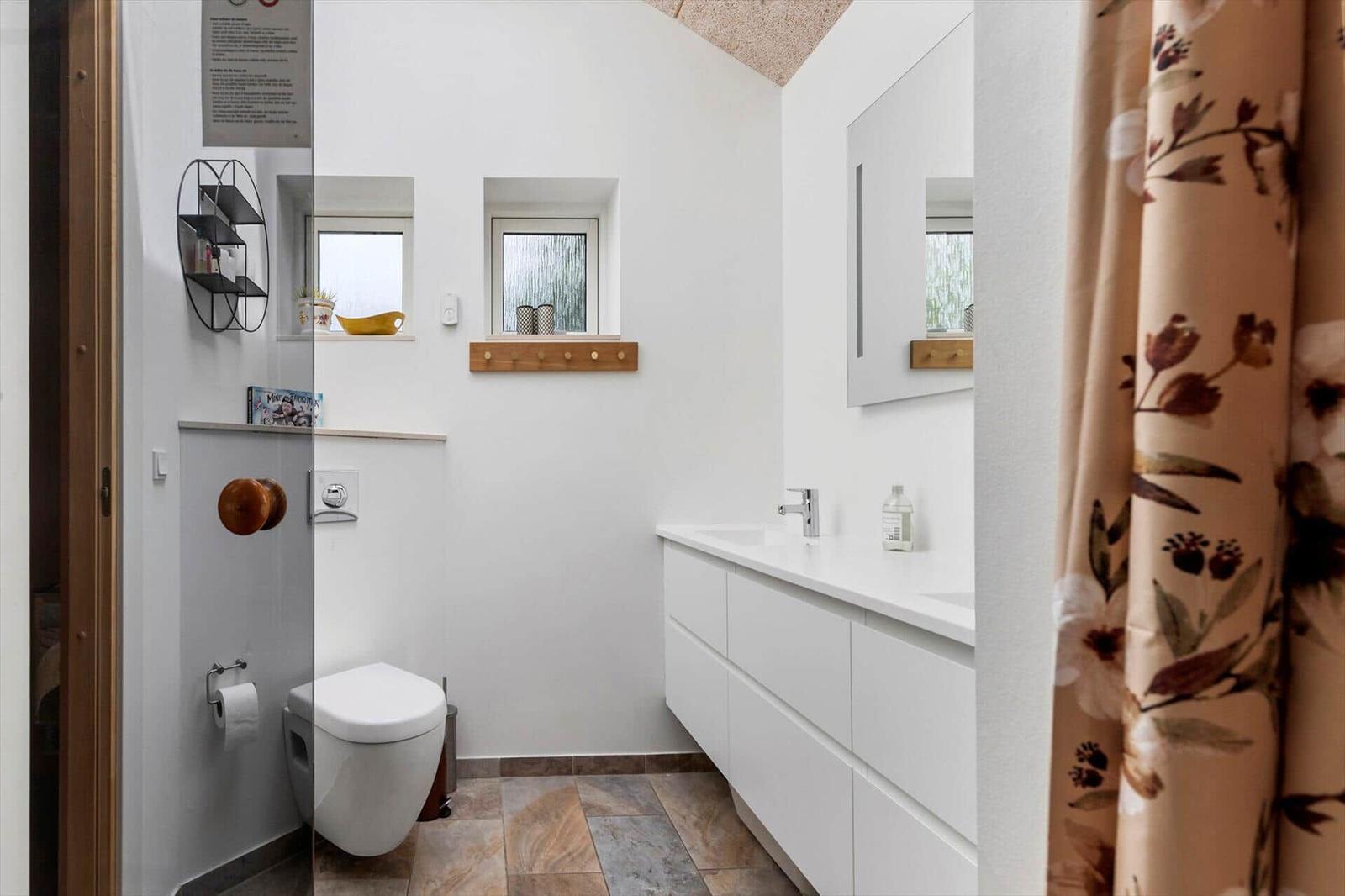 Badezimmer mit Toilette, Waschbecken und zwei Fenstern. Fliesenboden und Spiegelschrank.