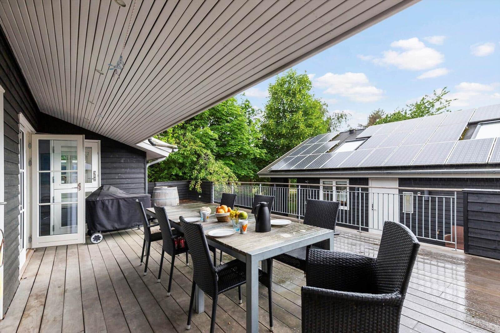 Terrasse mit Tisch, Stühlen und Grill unter Dach. Im Hintergrund Solaranlage auf Hausdach.
