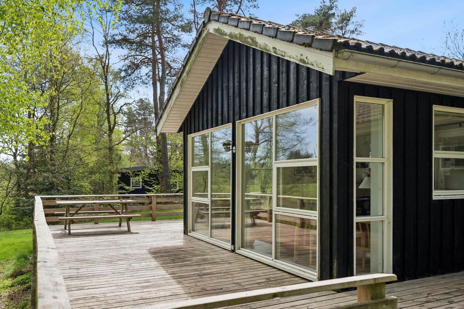 Schwarzes Haus mit Holzterrasse und Glasfront. Umgeben von Bäumen und Gras.