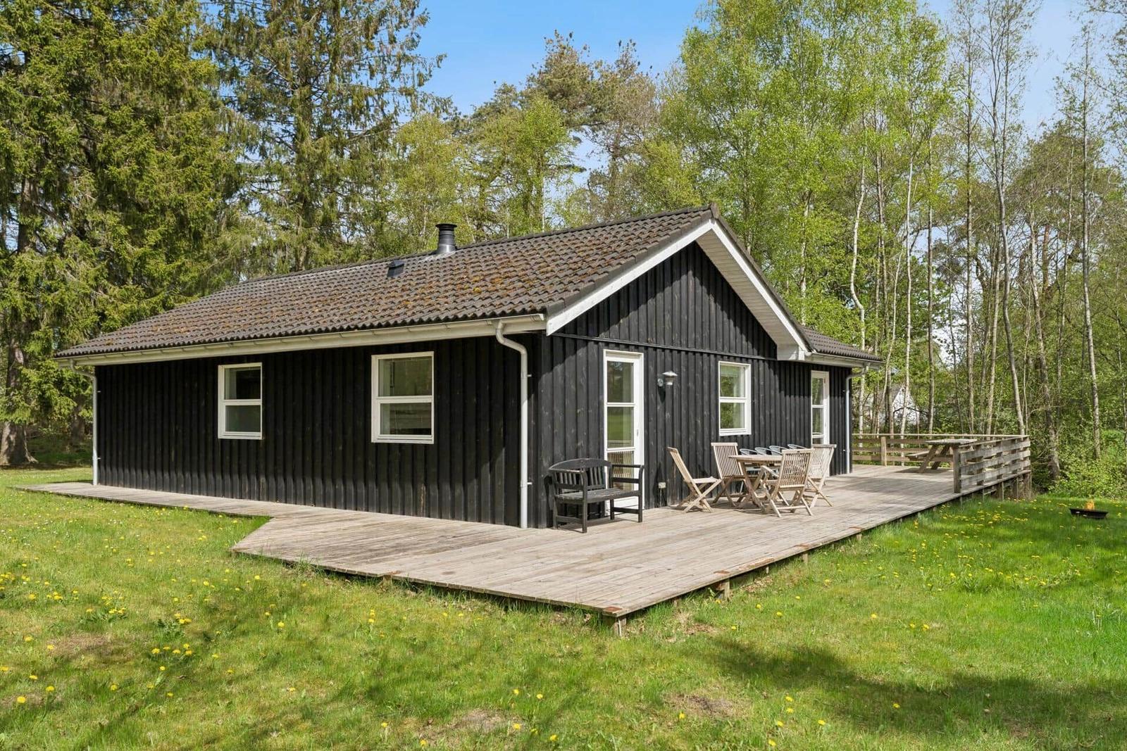 Schwarzes Holzhaus mit Terrasse und Garten. Holztisch, Stühle und Grill sind vorhanden.