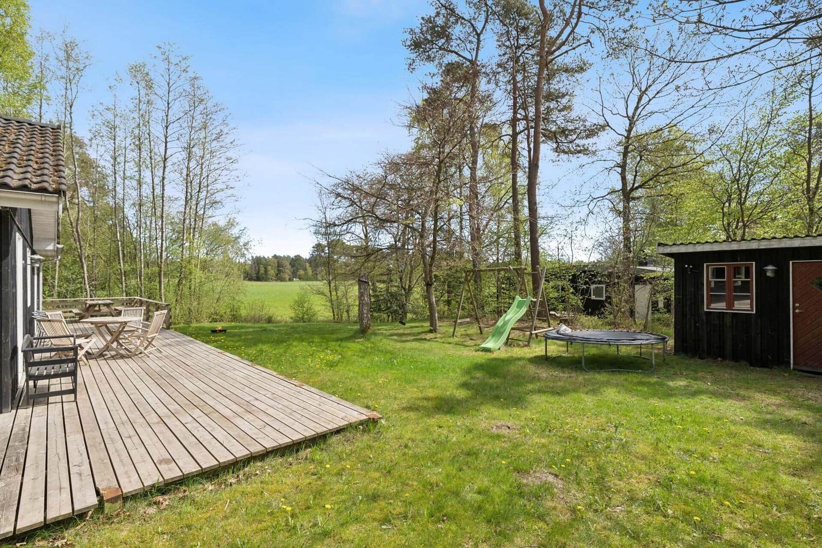 Hinterhof mit Terrasse, Trampolin und Schaukel. Grünfläche und Bäume umgeben die Hütte.