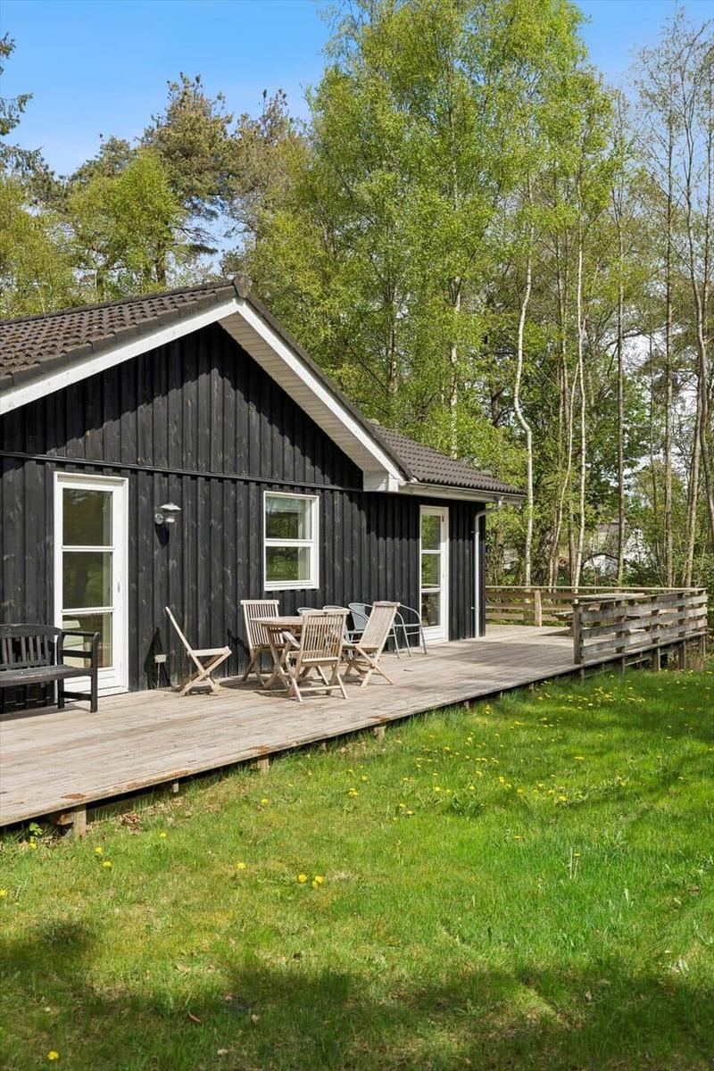 Schwarzes Holzhaus mit Terrasse, Garten und Bäumen im Hintergrund.