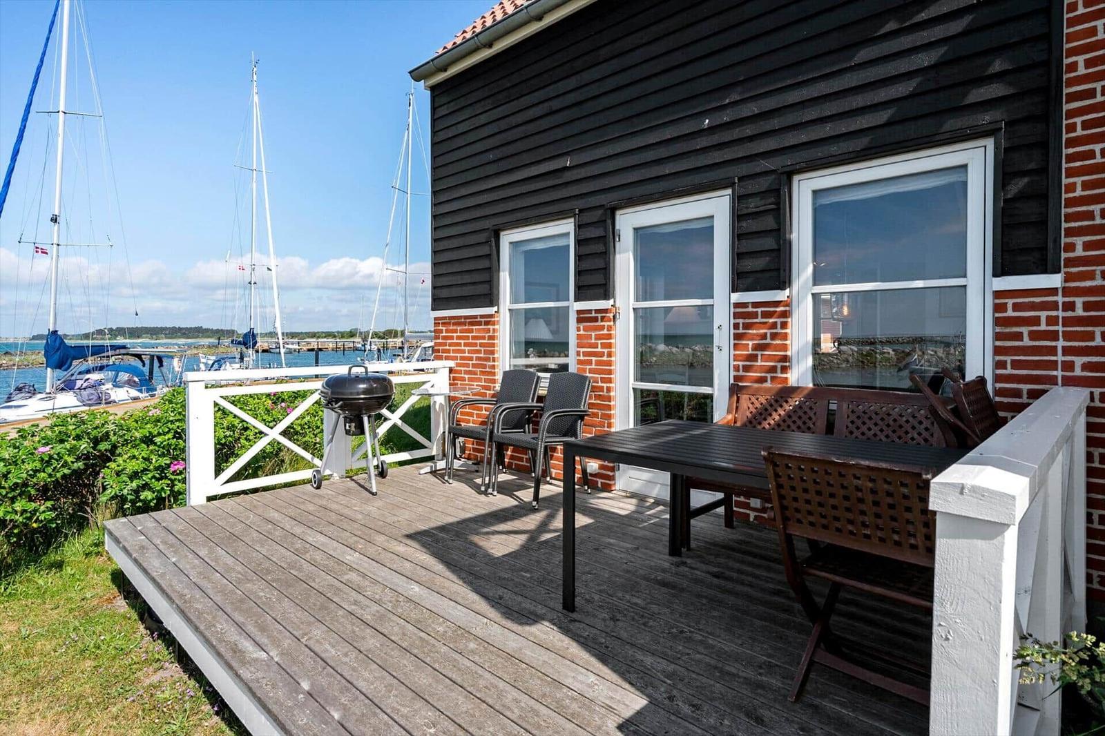 Terrasse mit Grill, Tisch und Stühlen. Blick auf Segelboote am Wasser.