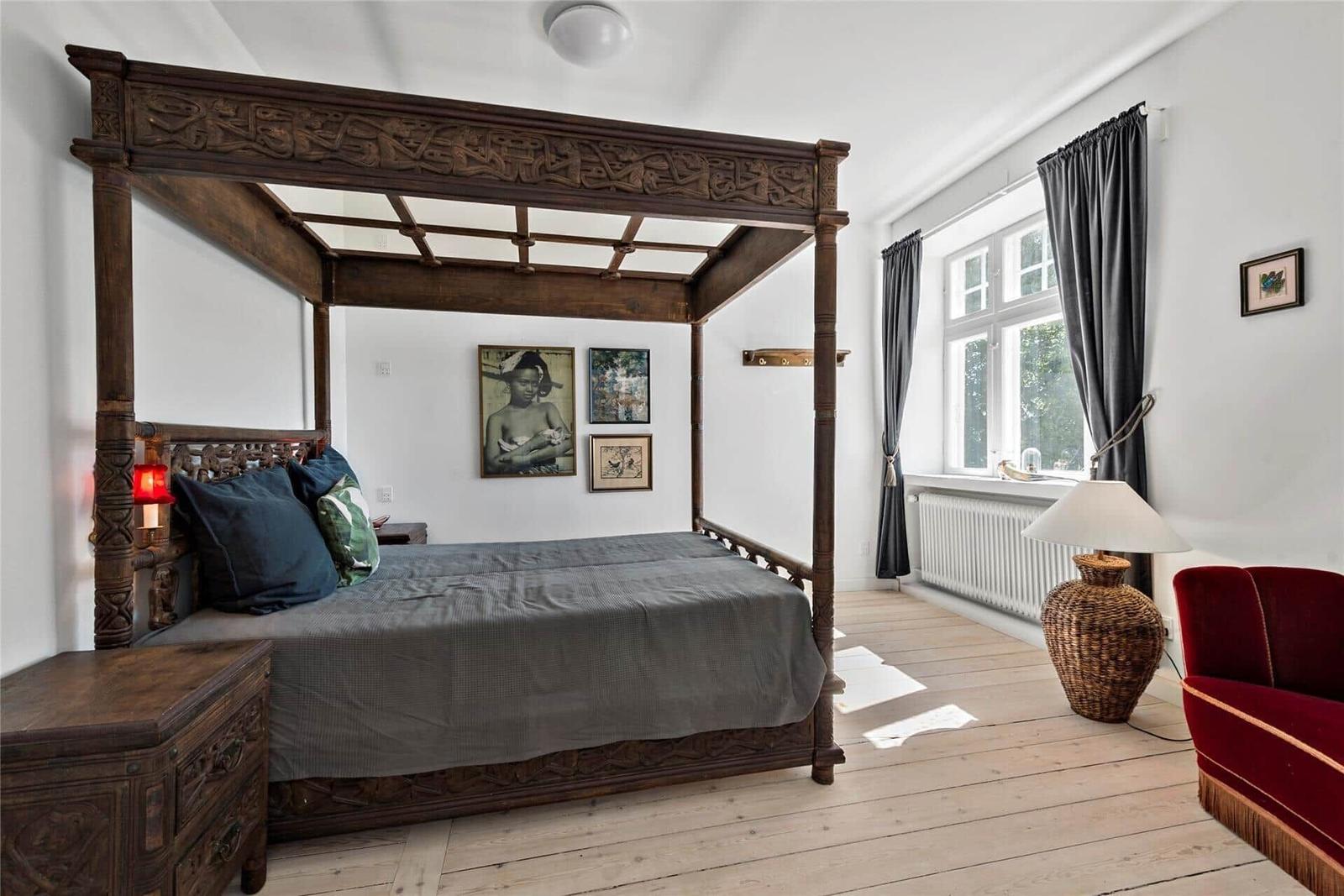 Schlafzimmer mit massivem Bett, Holzboden und Fenster mit Vorhängen.
