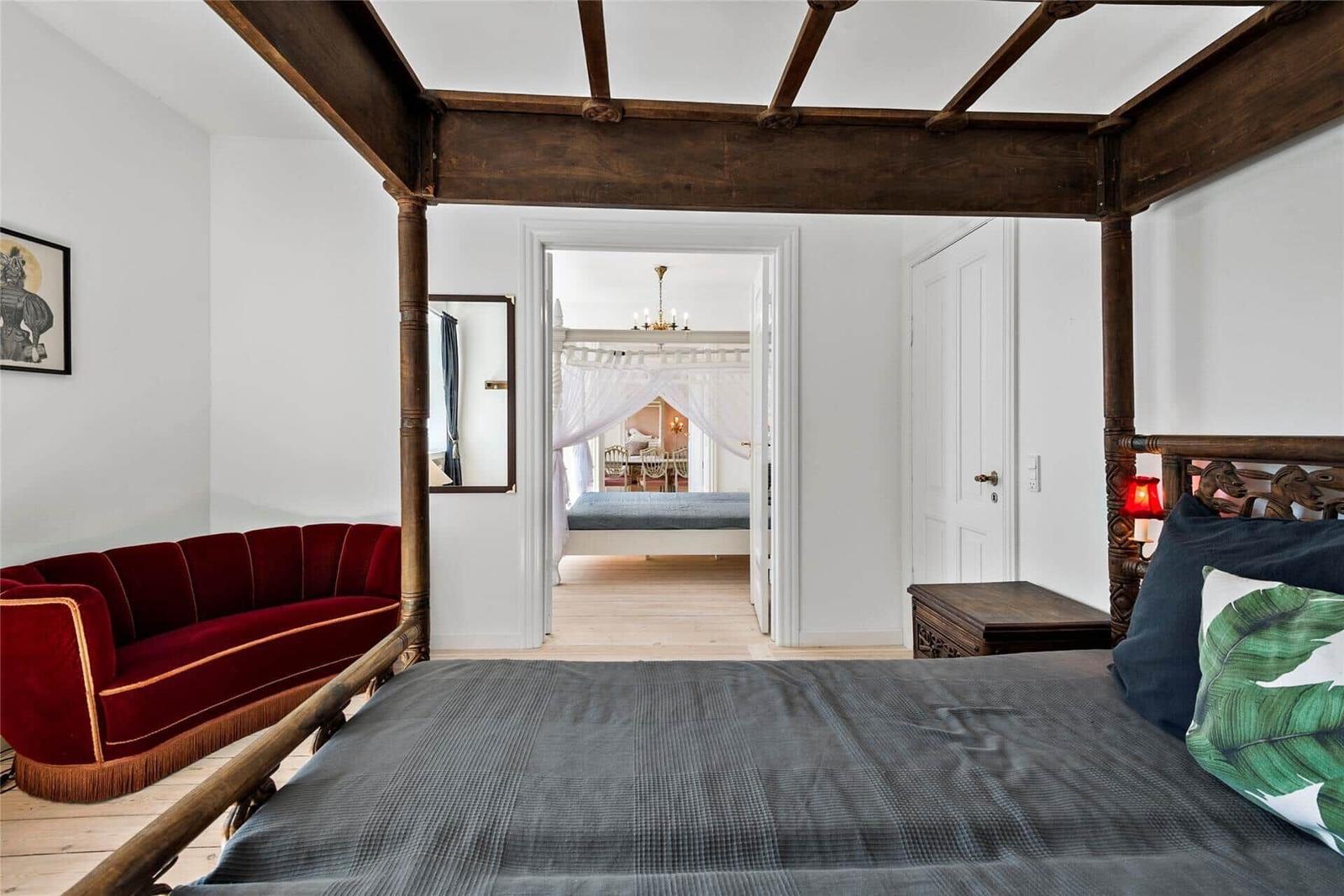 Schlafzimmer mit vierpostigen Bett, rotem Sofa und Blick in angrenzenden Raum.