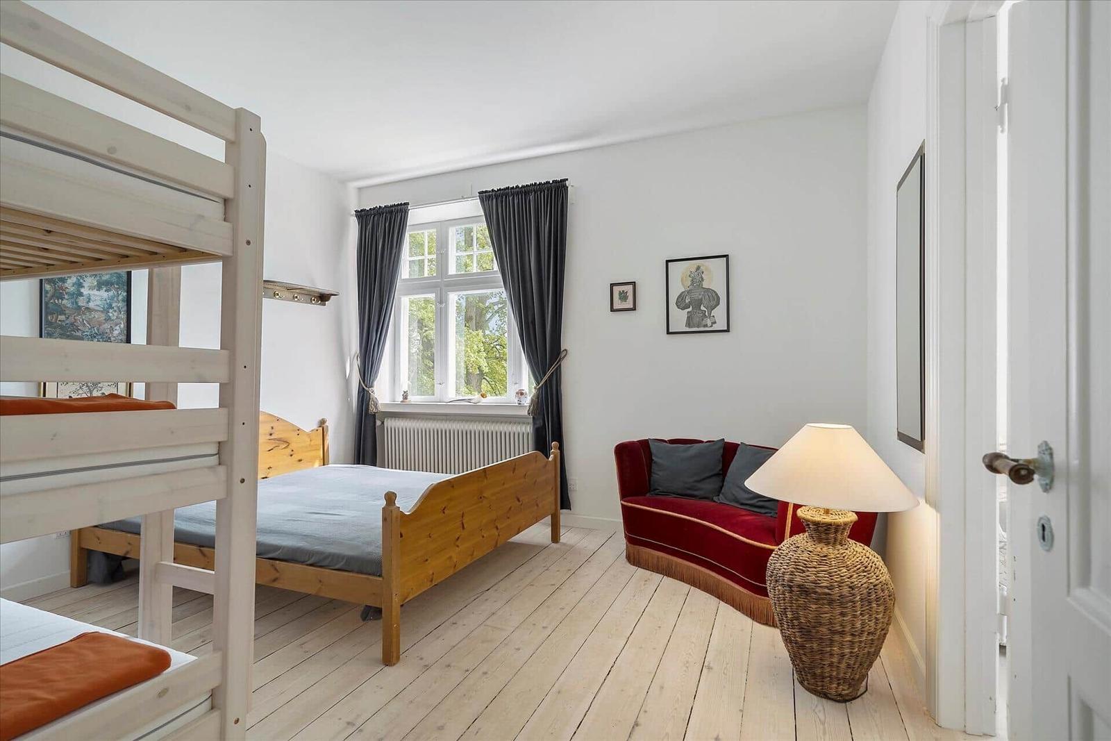 Zimmer mit Doppelbett, Etagenbett und rotem Sofa. Holzboden und Fenster mit Vorhängen.