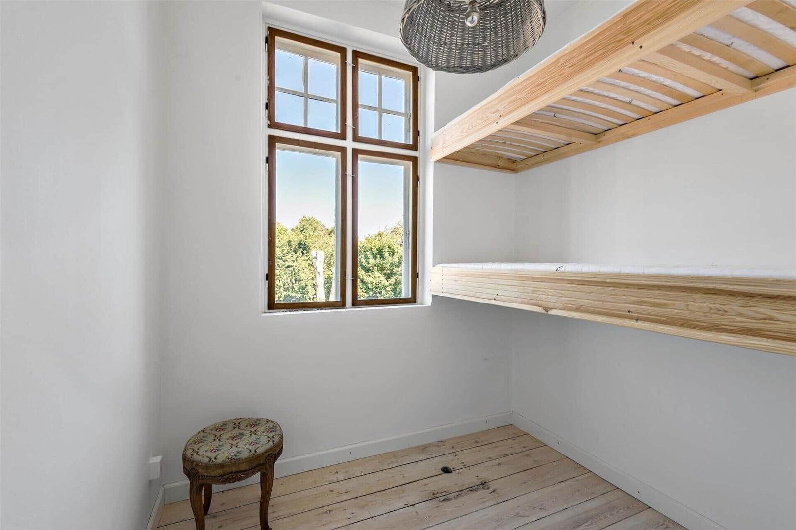 Ein Raum mit Holzboden, Fenster und erhöhtem Bett. Ein Stuhl steht daneben.
