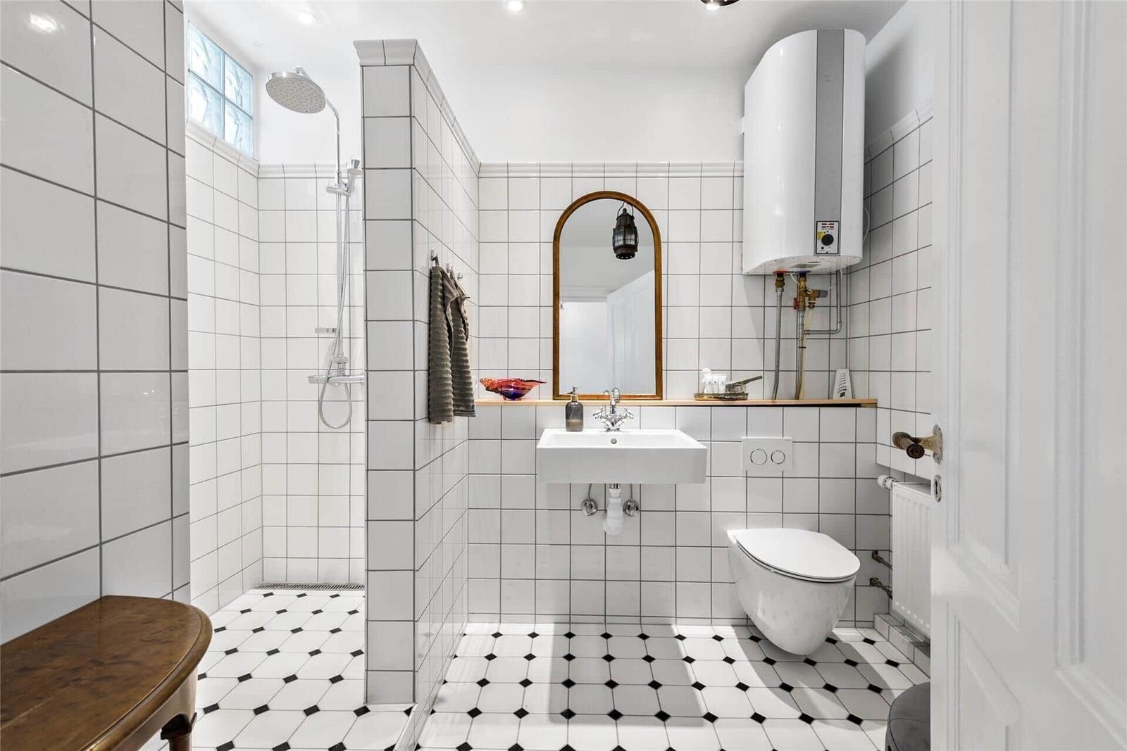 Badezimmer mit Dusche, Waschbecken und Toilette. Weiße Fliesen und schwarz-weiße Bodenfliesen.