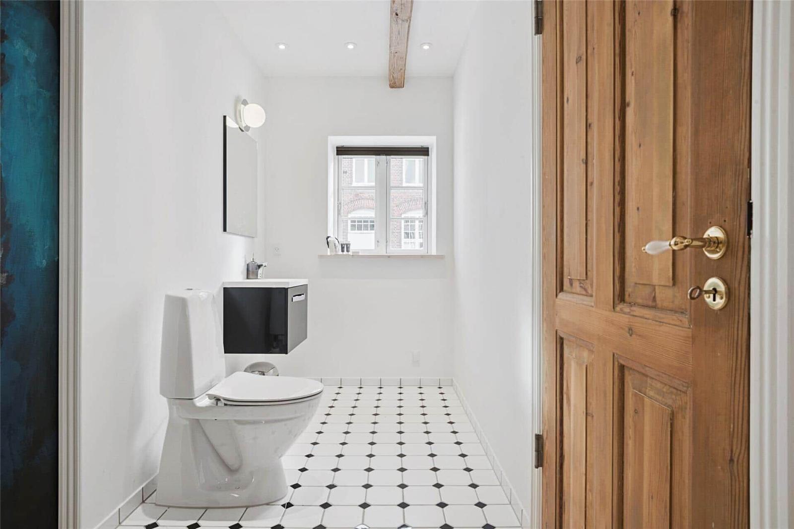 Badezimmer mit Toilette, Waschbecken und Fenster. Holztür und schwarz-weiße Fliesen.