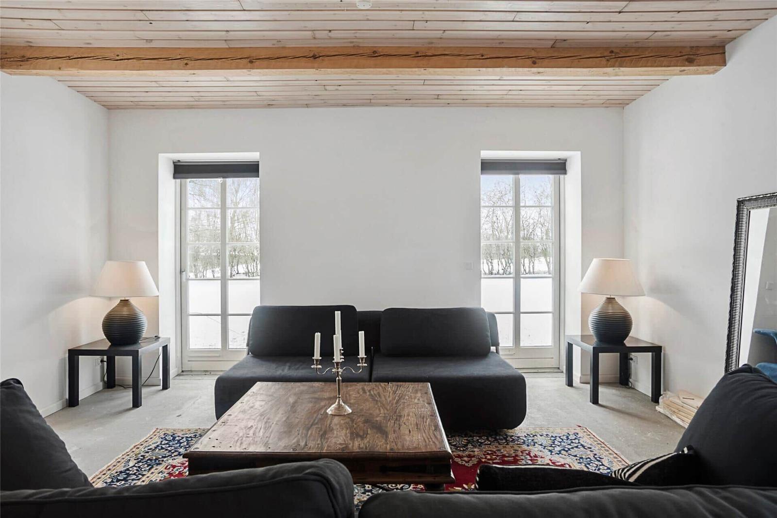 Wohnzimmer mit dunklem Sofa, Holztisch und Fenstern. Decke mit Holzbalken.