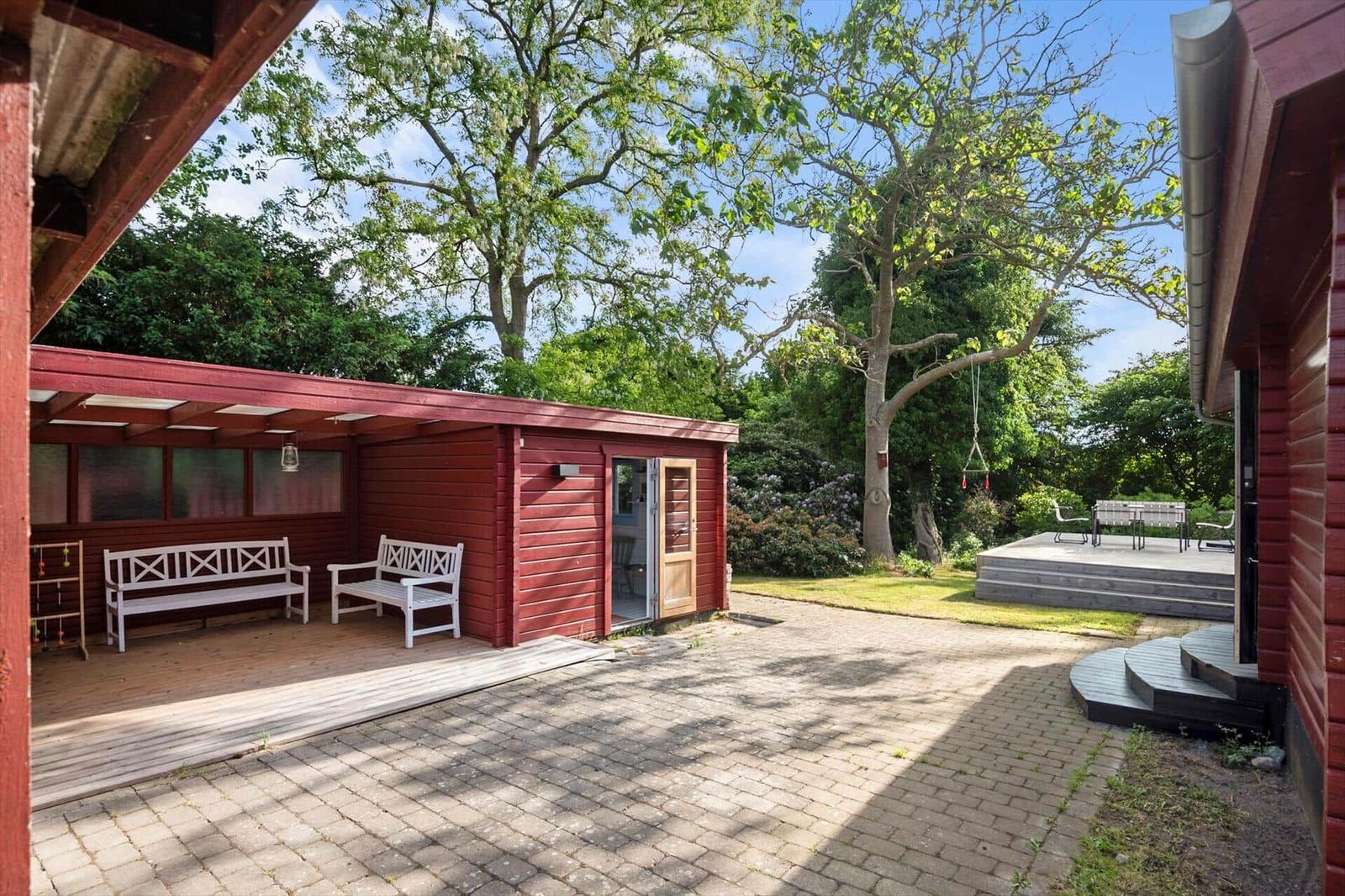 Roter Holzaufbau mit weißen Bänken und Terrasse unter Bäumen.