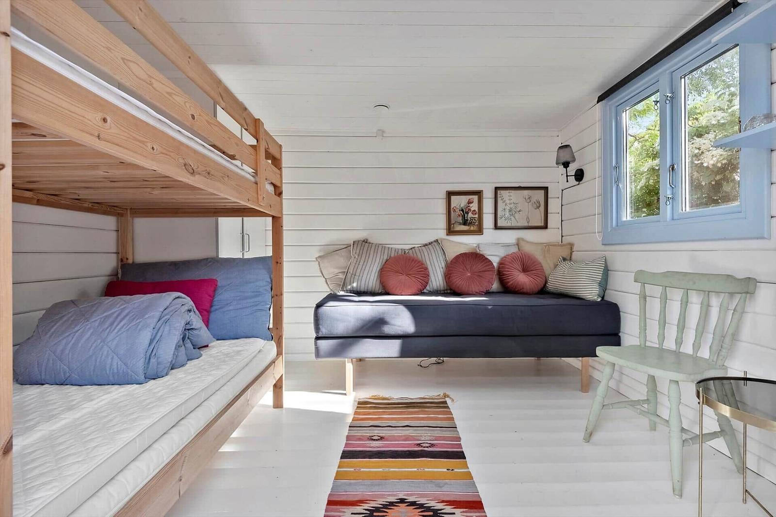 Zimmer mit Schlafcouch, Doppelstockbett und Holzstuhl. Fenster mit blauem Rahmen.