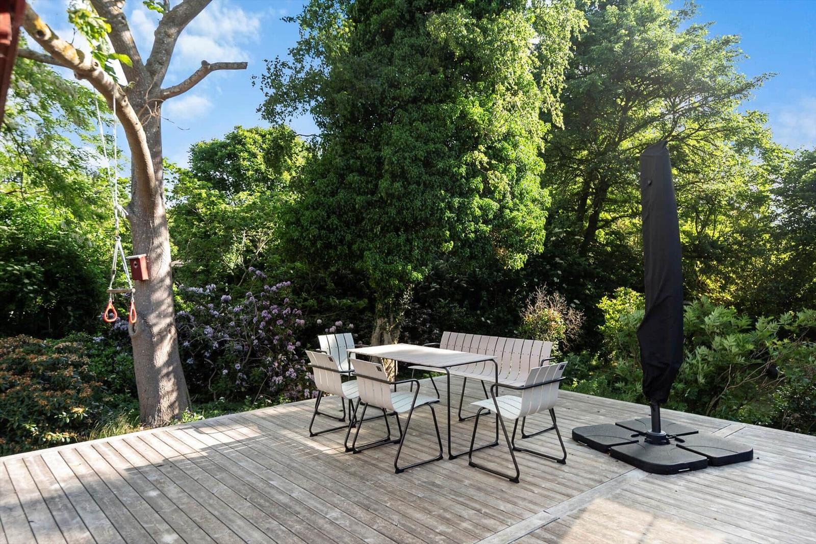 Holzterrasse mit Tisch und Stühlen, umgeben von Bäumen und Sträuchern. Ein geschlossener Sonnenschirm steht daneben.