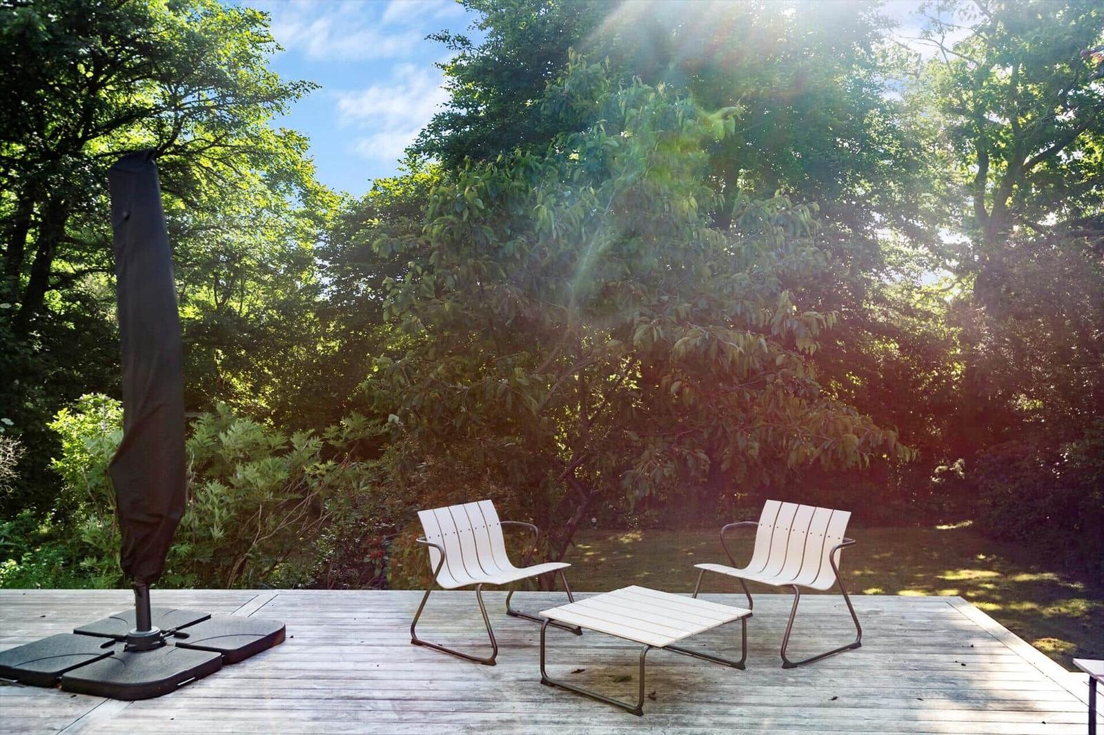 Terrasse mit zwei weißen Stühlen und Tisch, umgeben von Bäumen und Sonnenlicht.